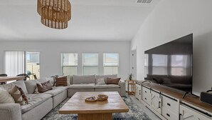 Ein modernes Wohnzimmer mit einer Eckcouch, einem Couchtisch, einem Fernseher und einem großen Fenster mit Jalousien.