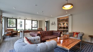 Ampla sala de estar com sofá de couro, uma poltrona cinza e mesa de centro de madeira. Há uma estante cheia de livros e objetos decorativos, e uma bicicleta fixada na parede.