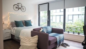 Quarto moderno com cama, sofá, manta de tricô, arte de parede com bicicleta e vista para o verde lá fora.