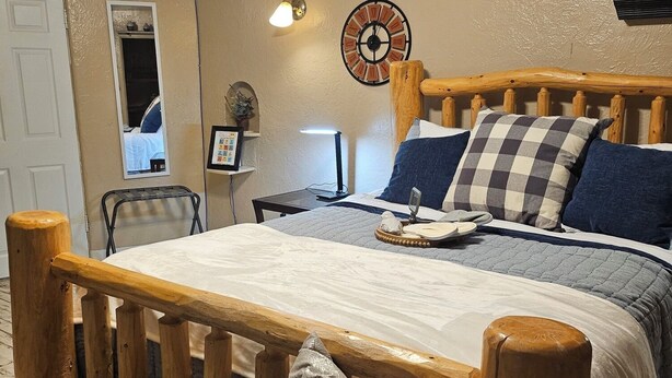 Un dormitorio acogedor con cabecera de cama de madera, una mesita de noche con lámpara, un reloj de pared y un estante pequeño con objetos decorativos.