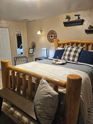 Un dormitorio acogedor con cabecera de cama de madera, una mesita de noche con lámpara, un reloj de pared y un estante pequeño con objetos decorativos.