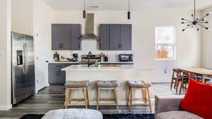 Une cuisine moderne avec des meubles gris, un îlot central et un coin repas avec des chaises en bois et un coussin rouge.