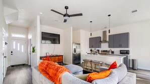 Un salon moderne avec un canapé gris, un plaid orange et une cuisine en arrière-plan.