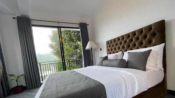 A large bed with a tufted headboard, grey and white bedding, a potted plant, and a view of greenery through a balcony. - The Lipton View Inn (Bandarawela)