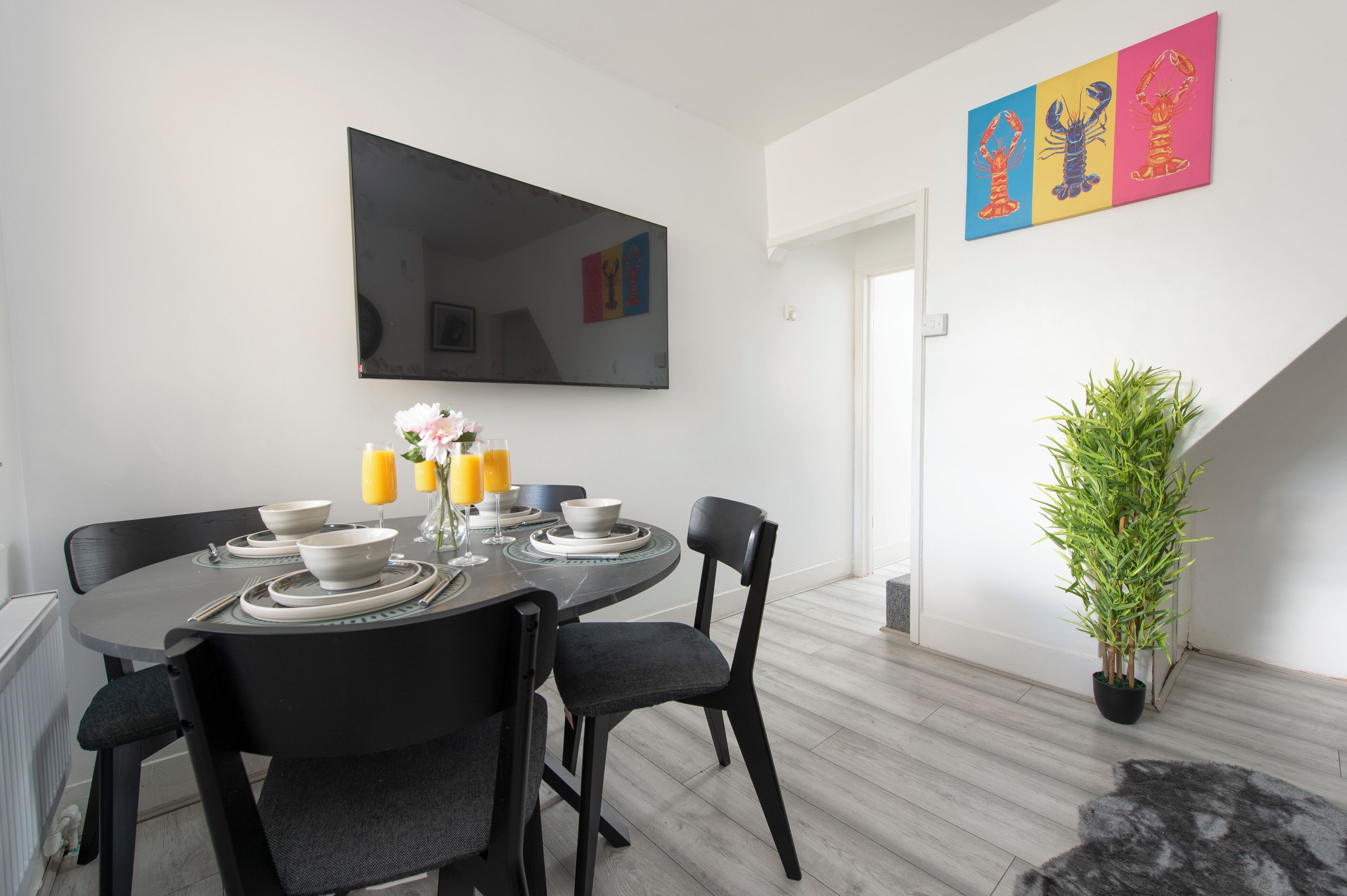 A dining area with a round table, black chairs, white plates, bowls, and a vase with flowers. There is a large mirror on the wall and a potted plant to the side.