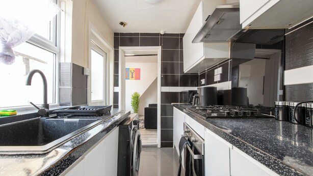 A modern kitchen with black countertops, stainless steel appliances, and a window with a curtain. - Stylish 4BR Home WiFi FreeParking (Thornton Heath)