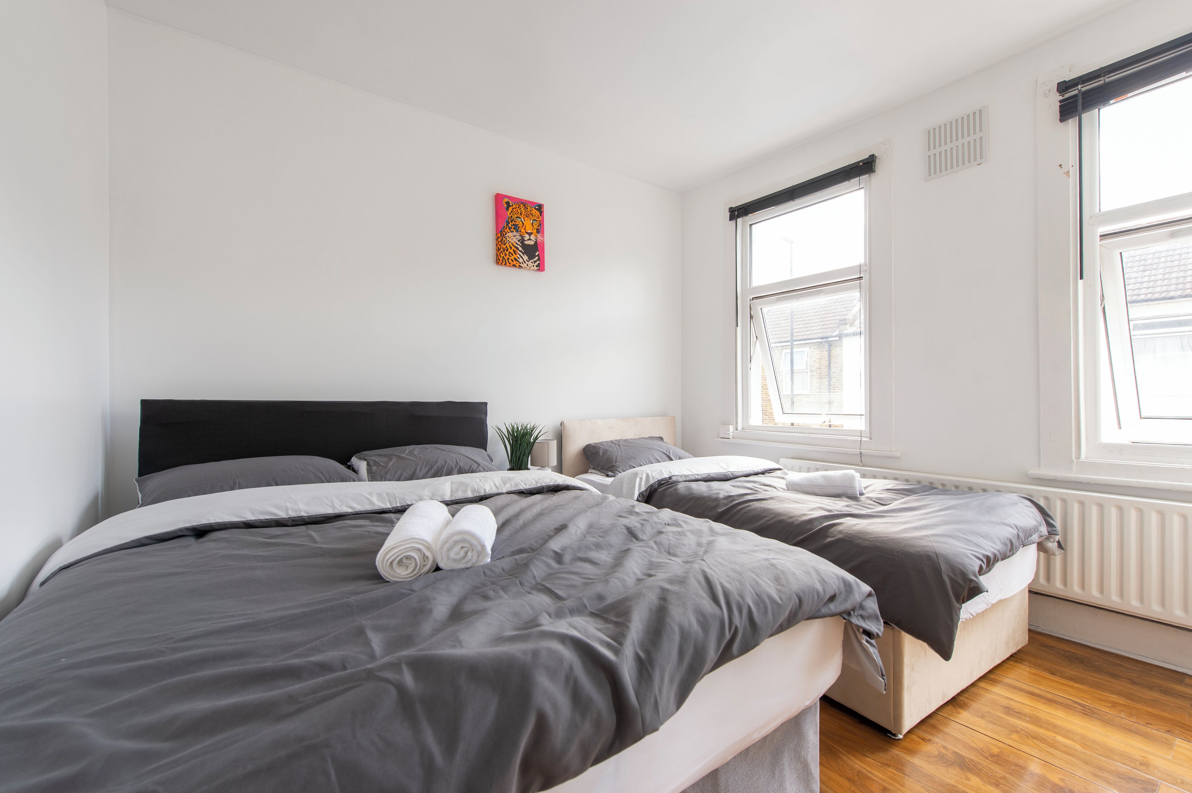 A modern bedroom with two beds, grey bedding, a colorful painting, and large windows.