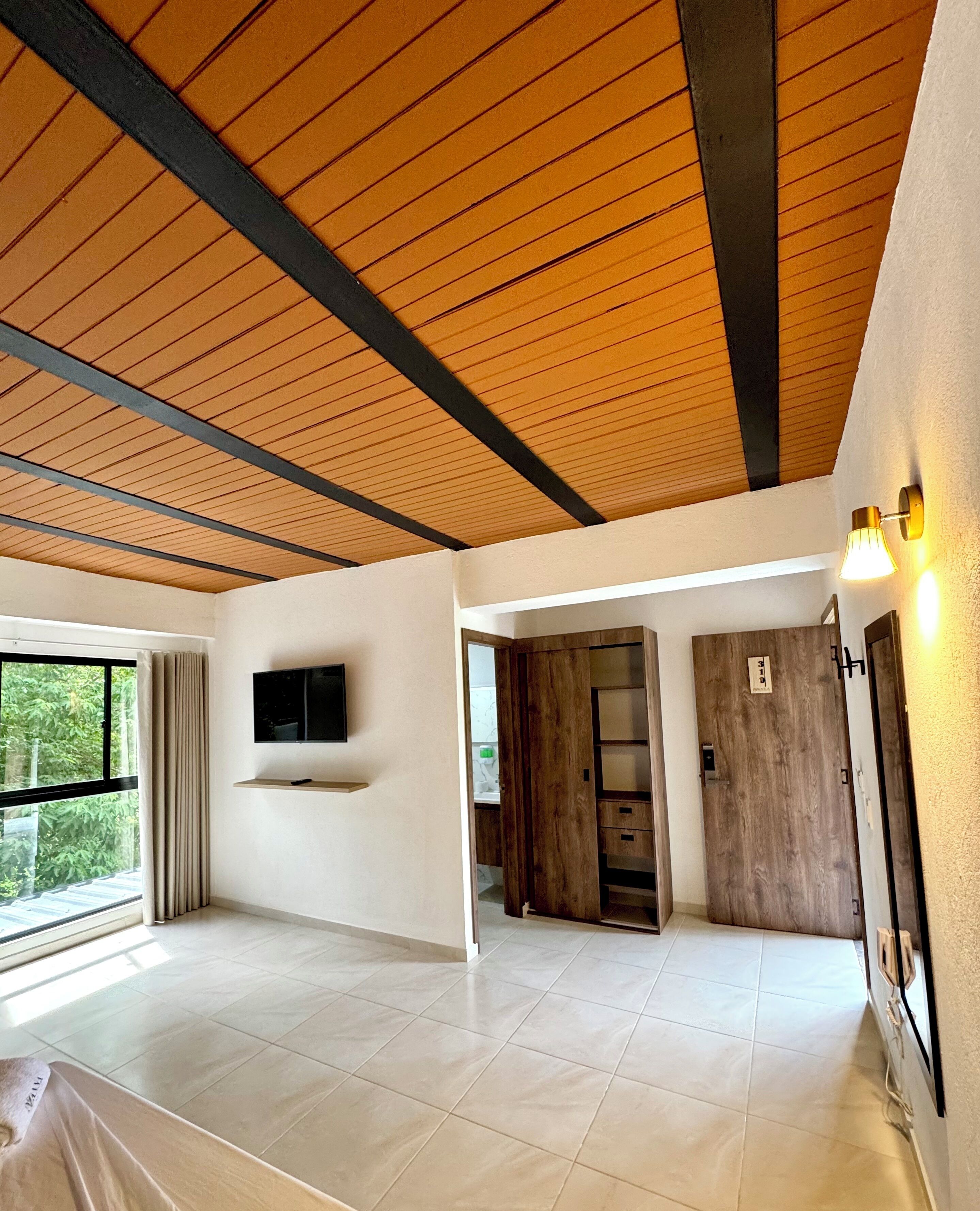 A spacious room with a high ceiling, a flat-screen TV mounted on the wall, and a large window with curtains.