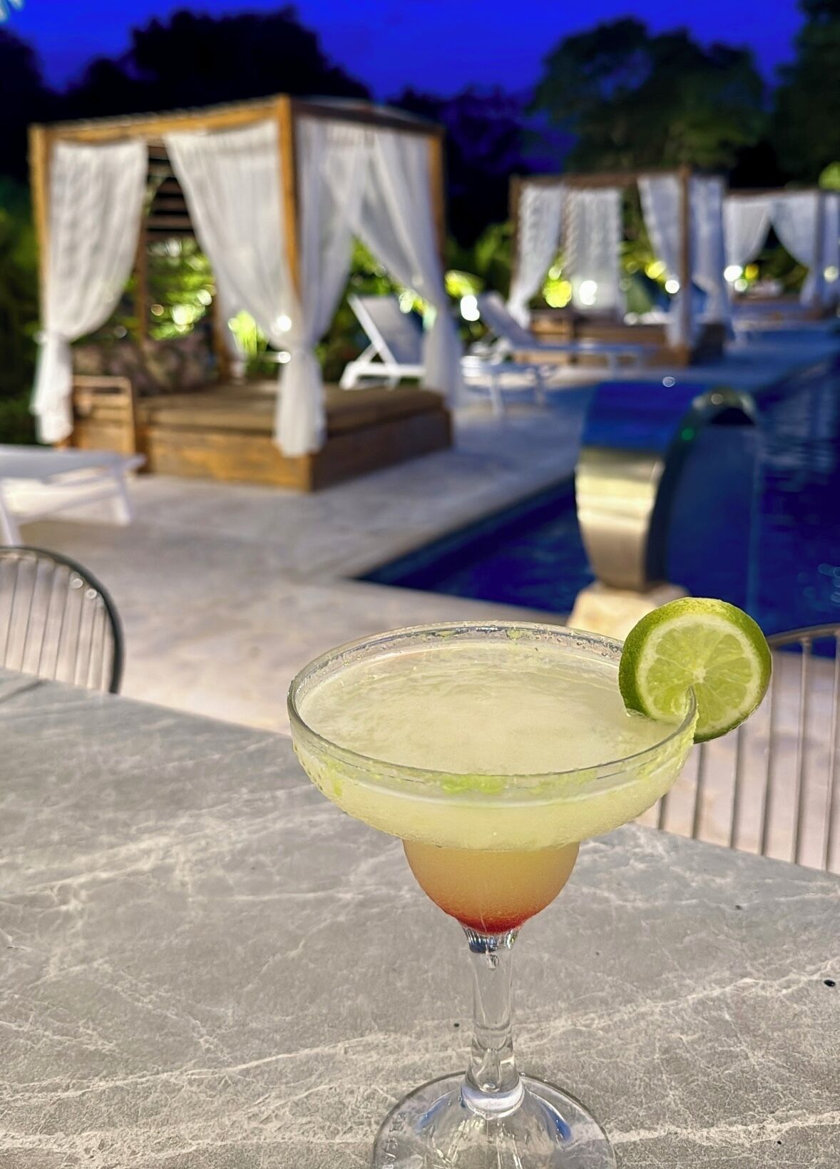 A cocktail on a table with a pool and lounge chairs in the background.