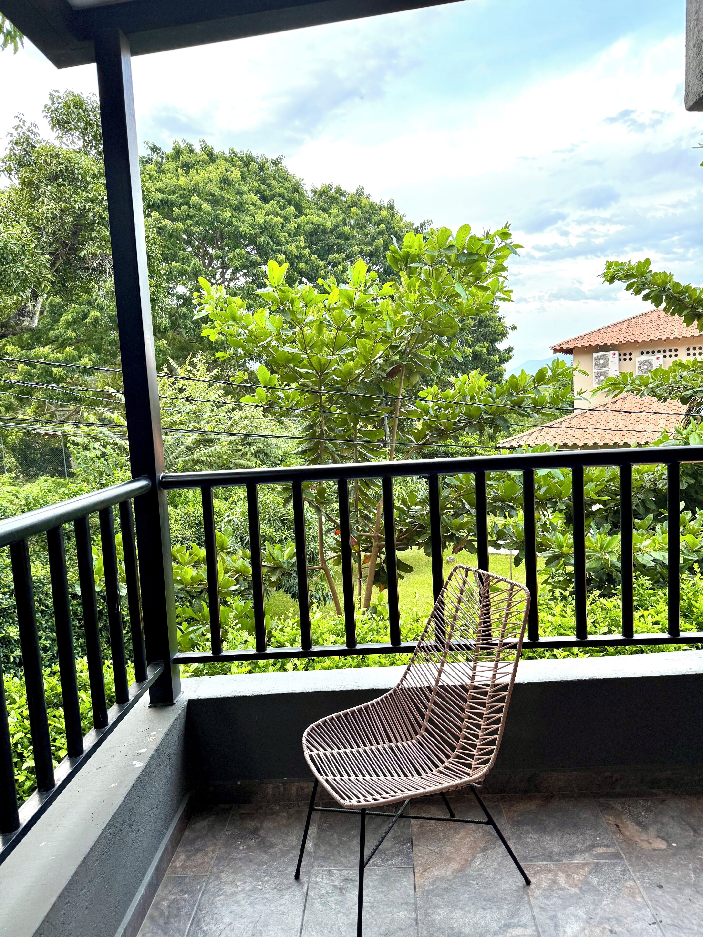 Un balcon avec une chaise, entouré d’une végétation luxuriante, et un bâtiment avec un toit en tuiles en arrière-plan.
