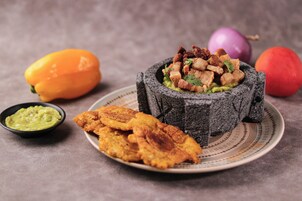 Un tazón de piedra lleno de un plato, acompañado de tajadas de plátano frito y un recipiente pequeño con salsa verde.