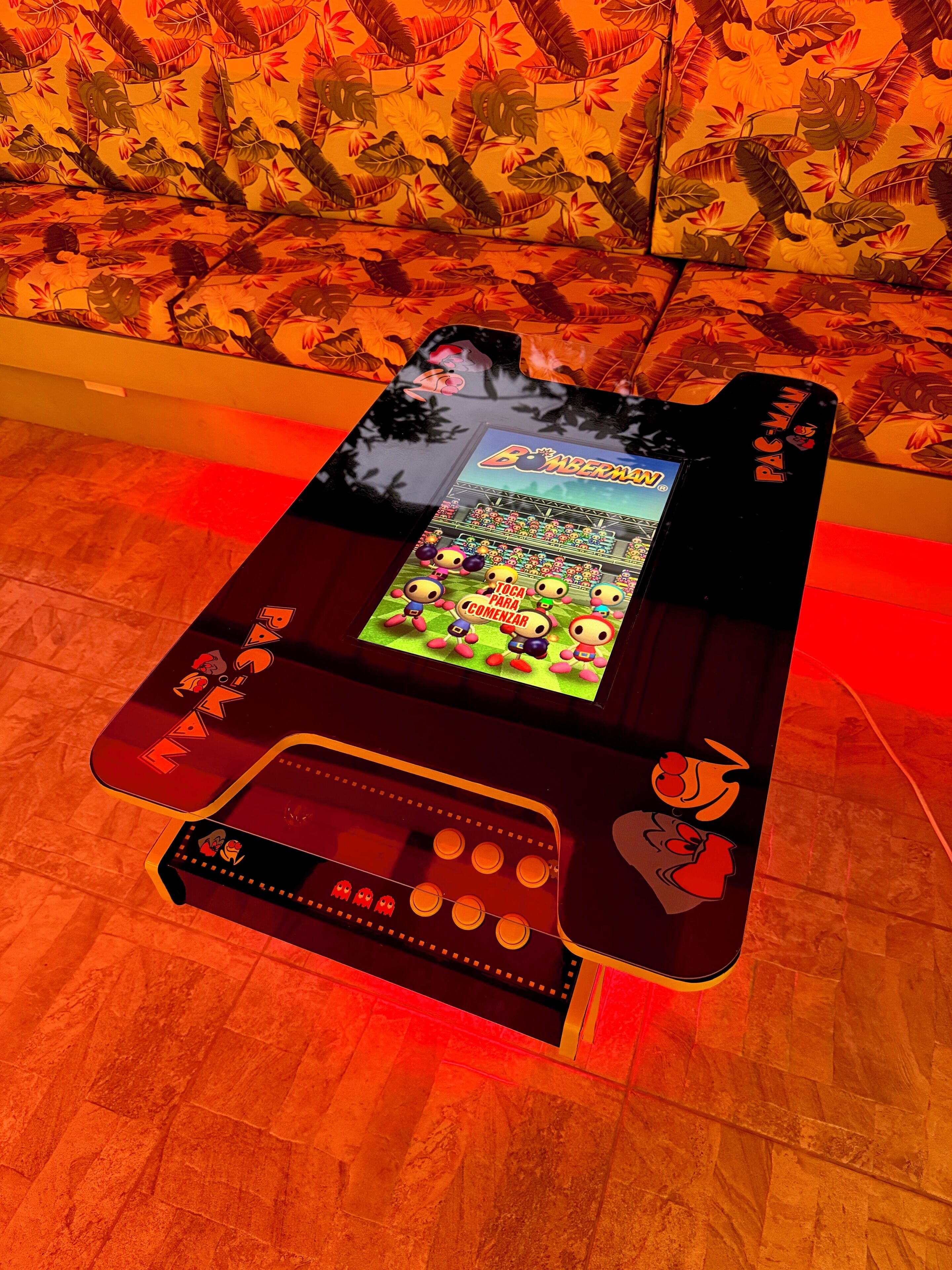 A Pac-Man arcade machine with a screen displaying a game, placed on a wooden floor with a tropical wallpaper background.
