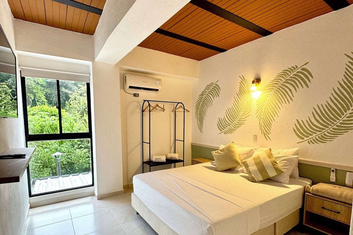 A bedroom with a large bed, a wardrobe, a hanging clothes rack, and a window with an air conditioning unit.
