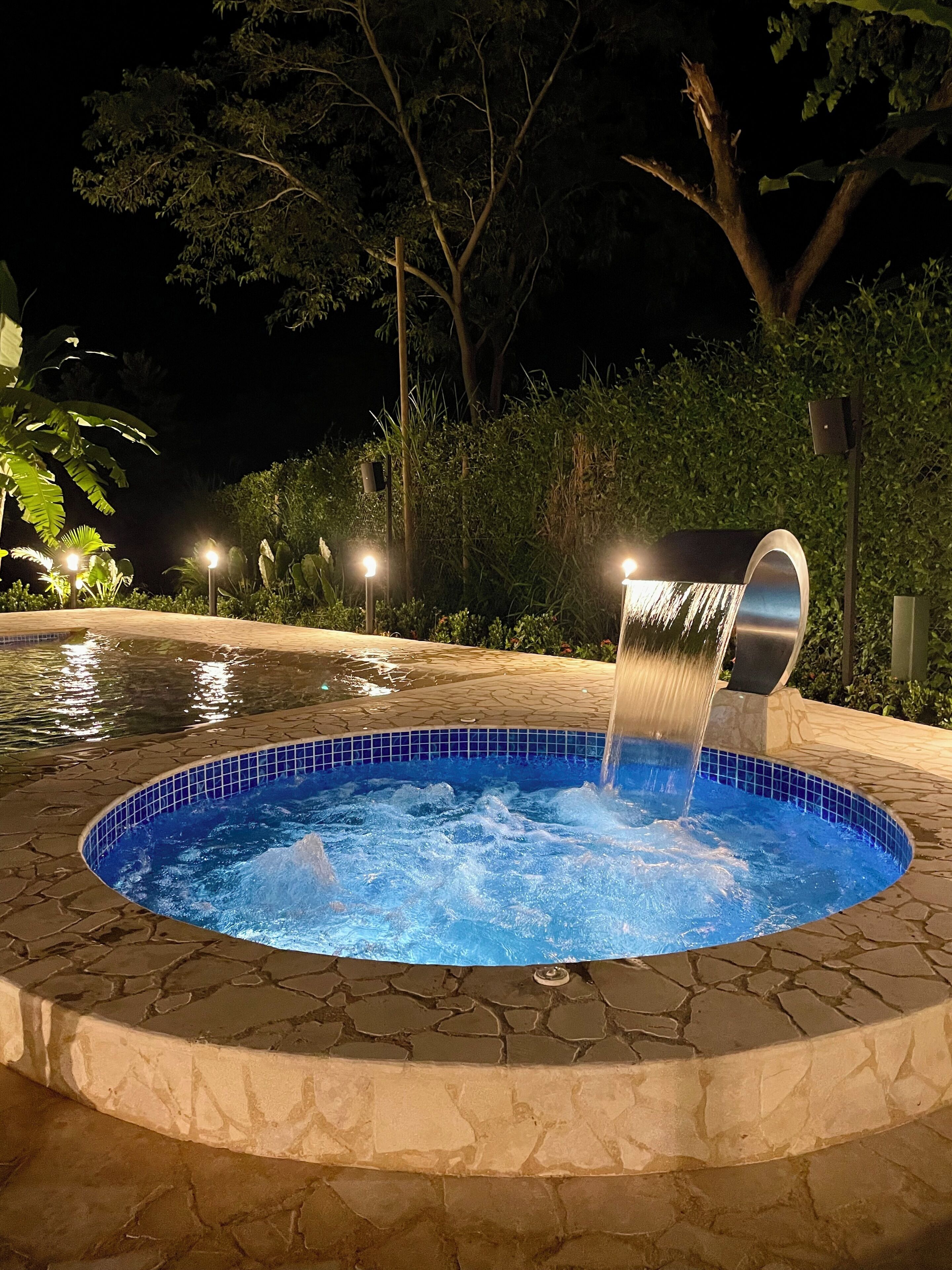 A hot tub with a waterfall feature, surrounded by a stone pool and illuminated by garden lights.