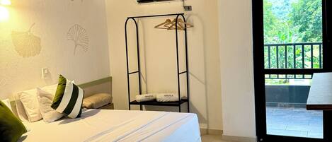 A bedroom with a bed, a wall-mounted air conditioner, a hanging clothes rack, and a view of greenery through an open door.