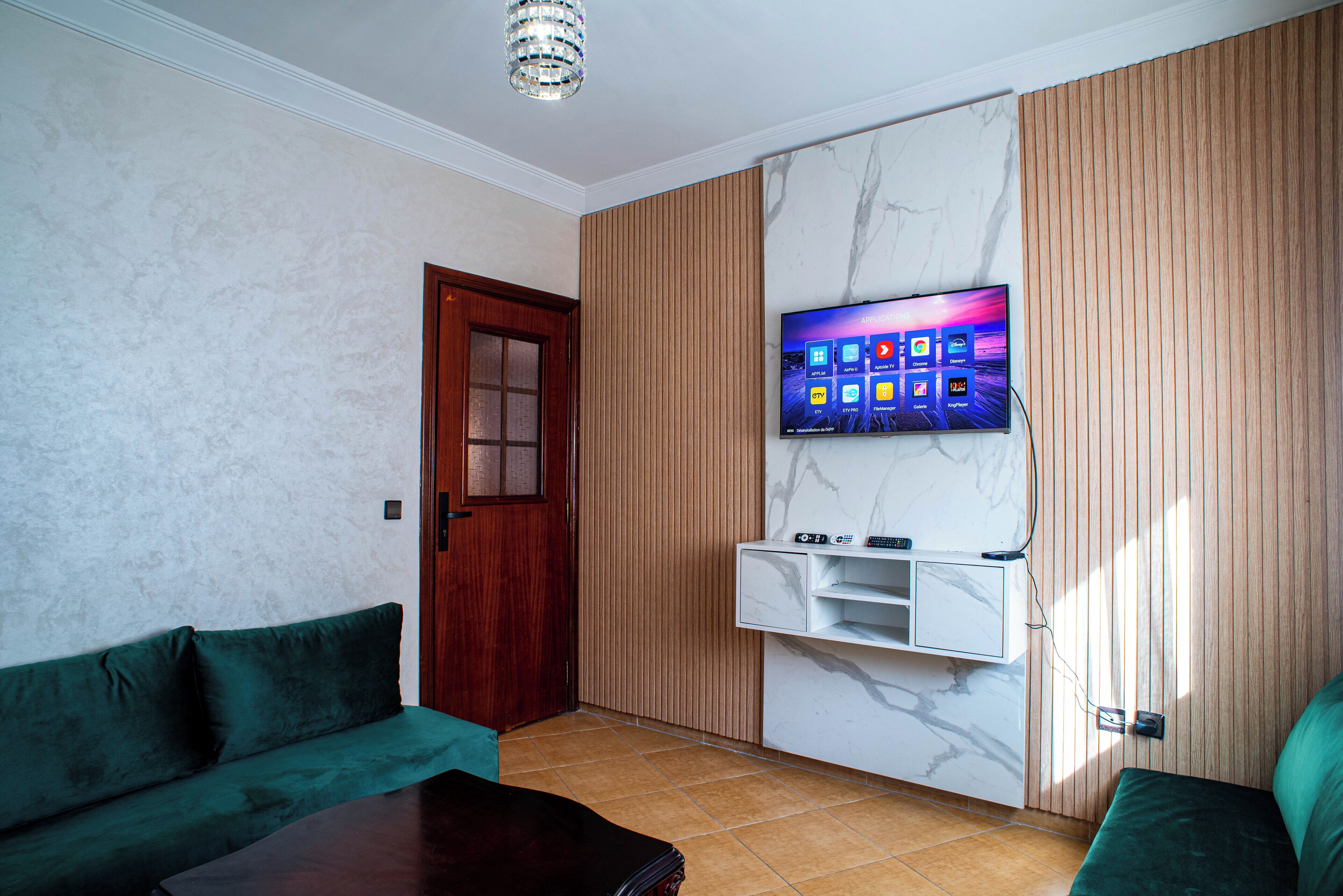 Un salón con un sofá verde, una puerta de madera, un televisor montado en una pared de mármol y un panel de madera a la derecha.