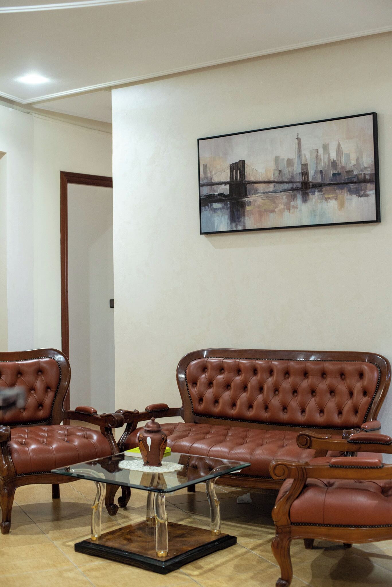 Un salon avec un ensemble de canapés en cuir marron, une table basse en verre et un tableau représentant un paysage urbain accroché au mur.