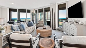 A spacious living room with a large window overlooking a cityscape, a comfortable sofa set, a round woven coffee table, and a flat-screen TV mounted on the wall. - Portofino Island Resort 4-704 (Pensacola Beach)