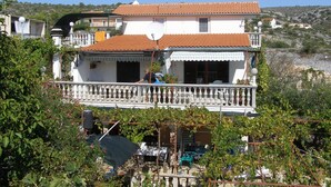 Ein zweistöckiges Haus mit Balkon, umgeben von Grünflächen und einer hügeligen Landschaft.