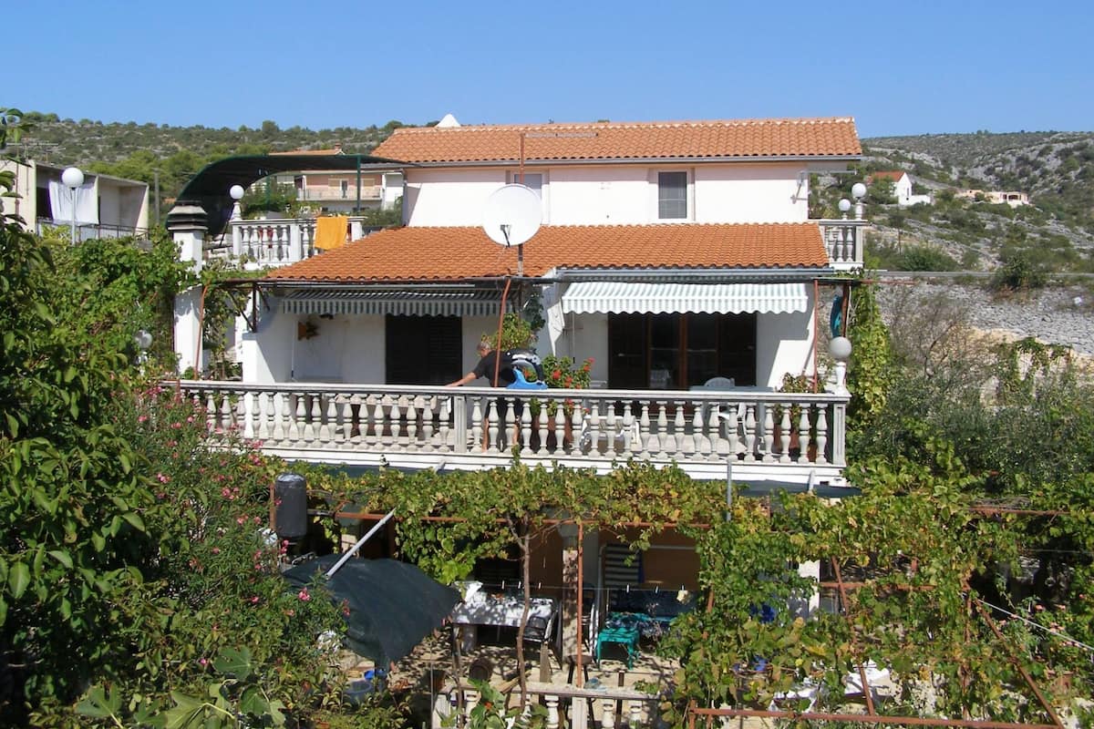 A two-story house with a balcony, surrounded by greenery and a hilly landscape.