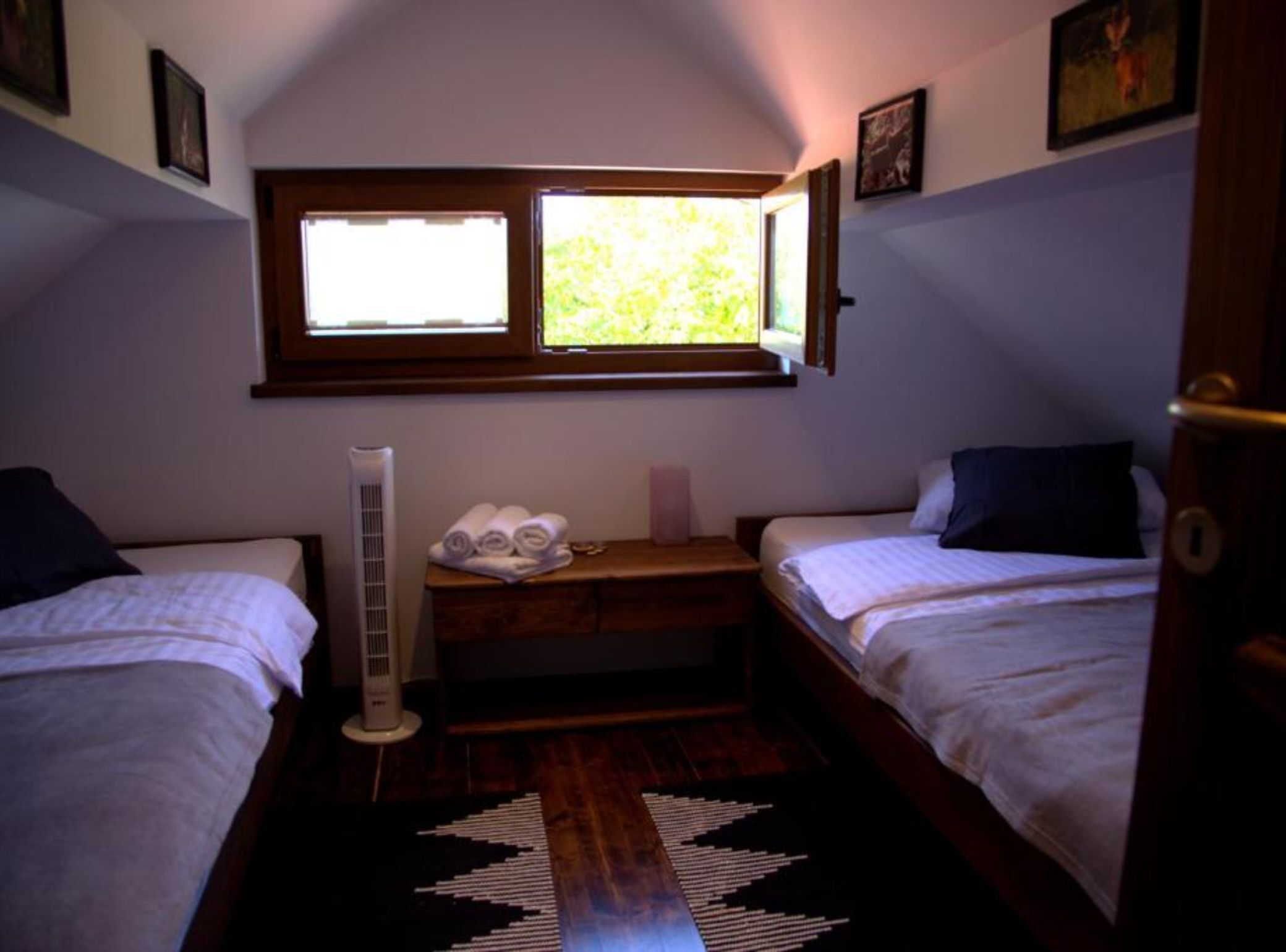 A small, dimly lit room with two single beds, a radiator, a window with a view of greenery, and a wooden bench with towels.