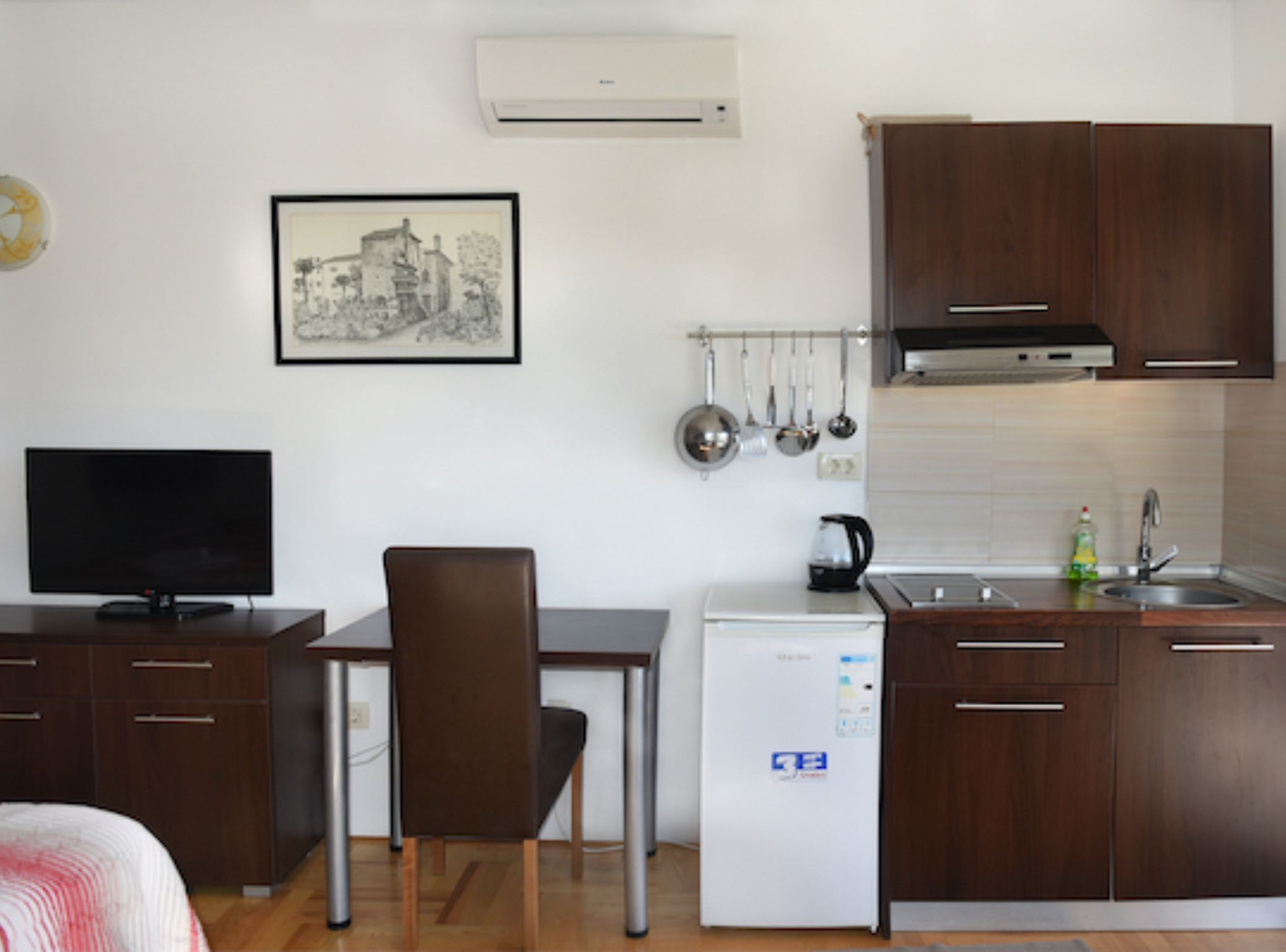 A compact kitchen with a refrigerator, stove, and sink. There's a dining table with a chair, a TV on a cabinet, and a framed picture on the wall.