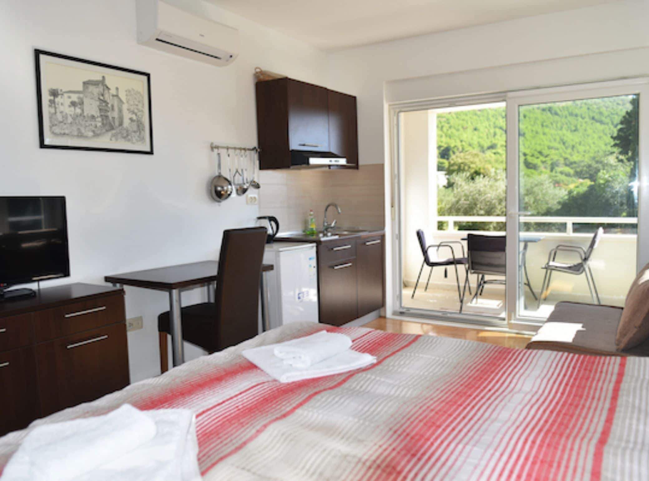 Habitación de hotel con cama, escritorio, kitchenette y balcón con vistas a la vegetación.