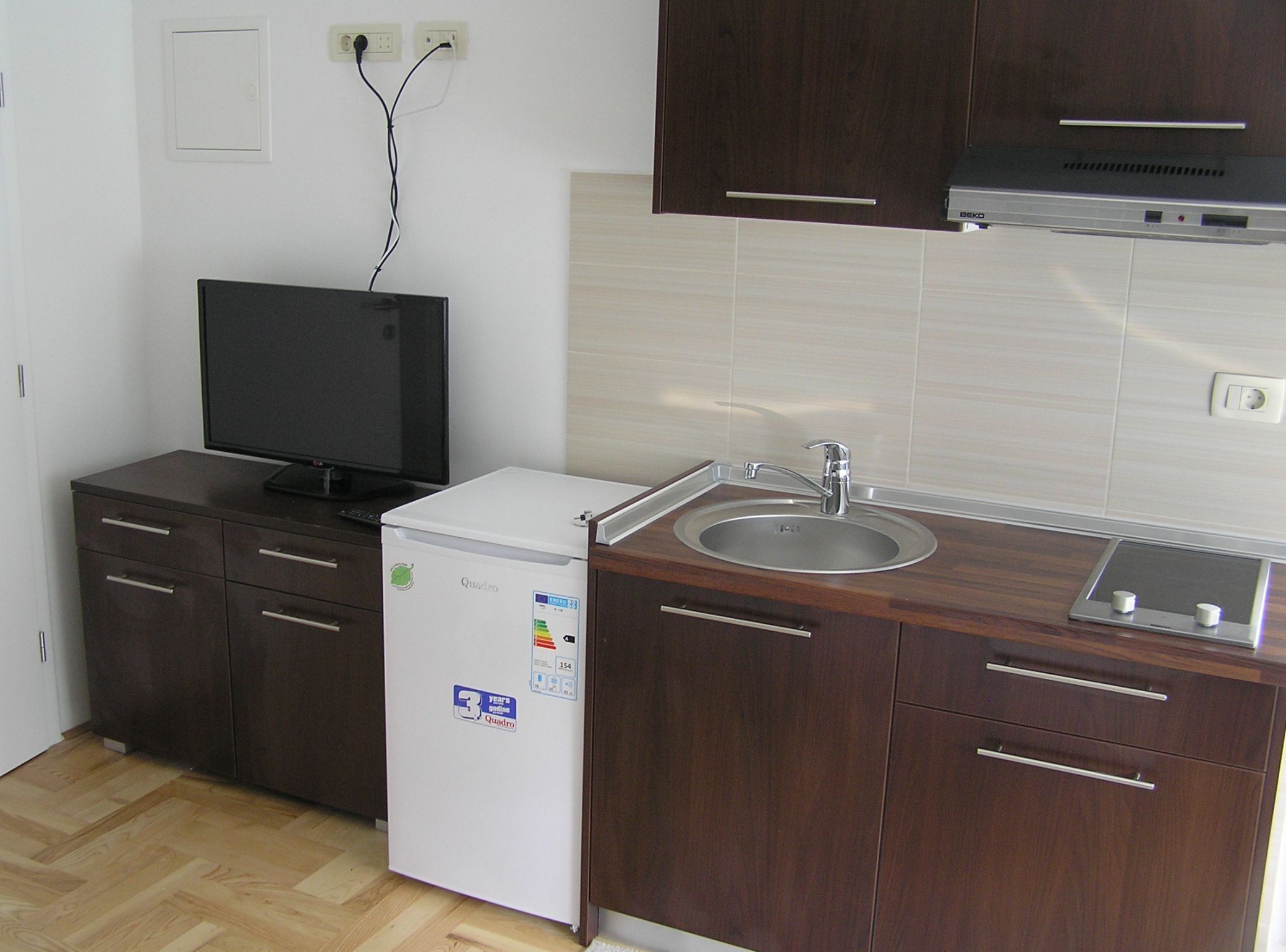 A compact kitchen with a refrigerator, a sink, a stove, and a television.
