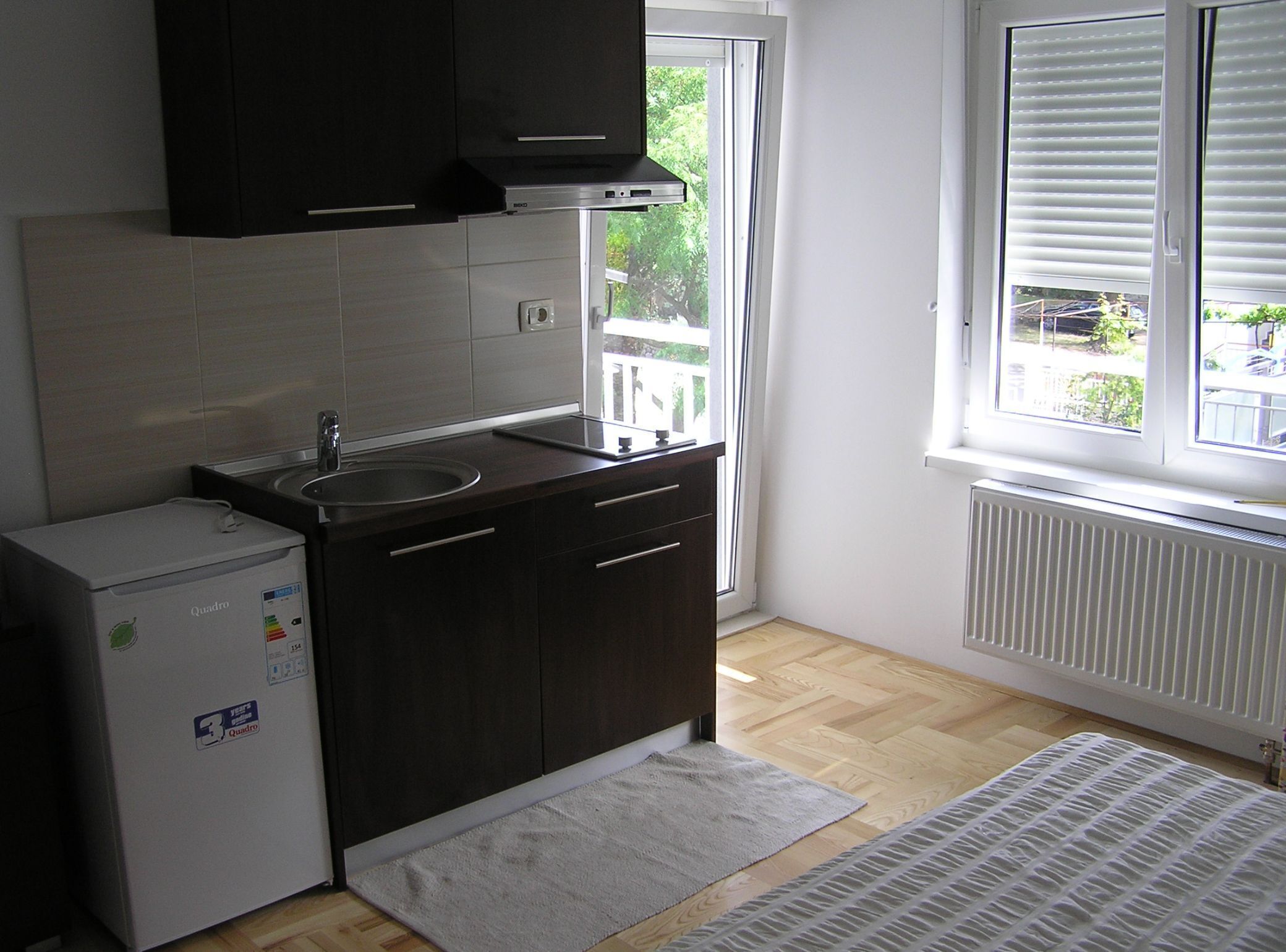Una cocina compacta con muebles oscuros, un refrigerador blanco y una ventana con persianas.