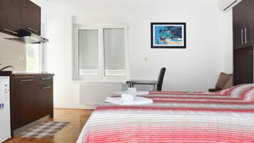 A compact hotel room with a bed, a small desk, a chair, a radiator, and a window with blinds.