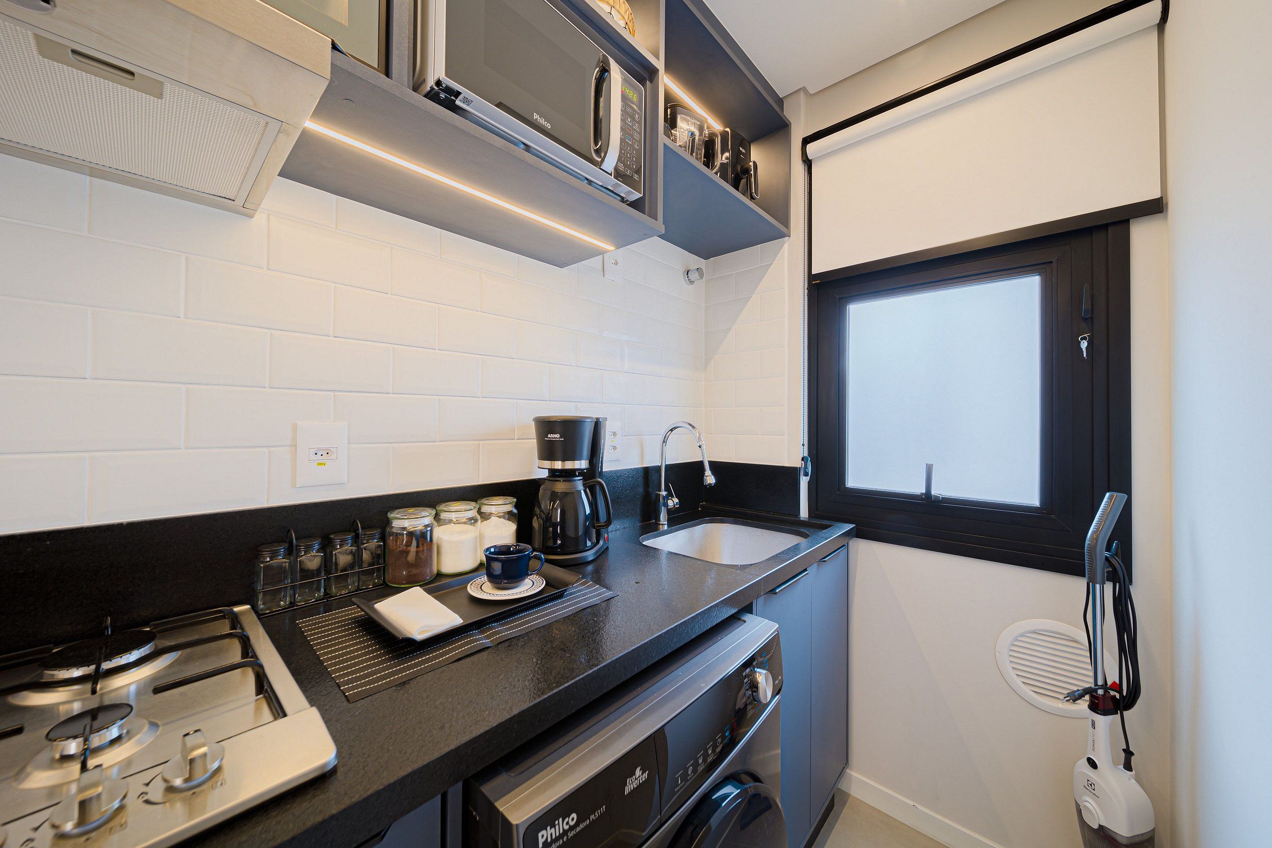 Una cocina compacta con estufa a gas, cafetera, pileta y ventana.