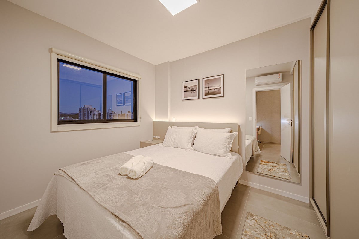 Une chambre avec une grande fenêtre, un lit recouvert d’une courtepointe blanche, deux tableaux encadrés au mur et un miroir.