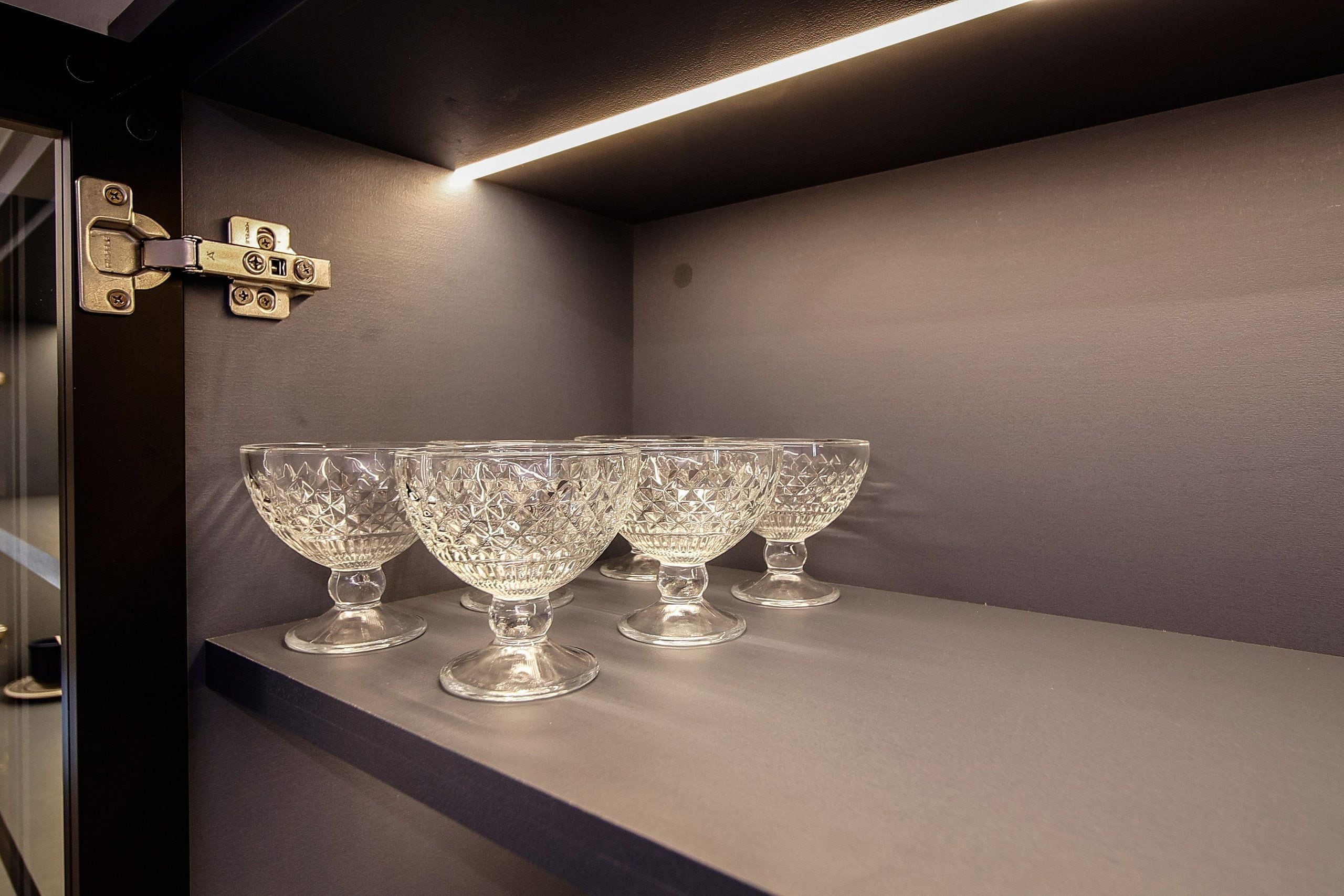 Un conjunto de cuatro copas de cristal transparente sobre una estantería, con un moderno aplique de luz encima.