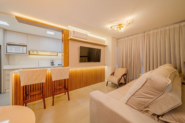 A modern living room with a sofa, a dining area with a table and chairs, a TV mounted on the wall, and an air conditioning unit. - Mingus Residencial Flipchave MIN000 (Joinville)