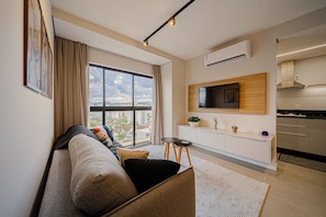 A modern living room with a sofa, a small table, a TV mounted on the wall, and a kitchen area with cabinets and a stove. - Mingus Residencial Flipchave MIN000 (Joinville)