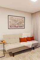 Uma sala de estar com sofá, uma obra de arte emoldurada e uma janela com cortinas.