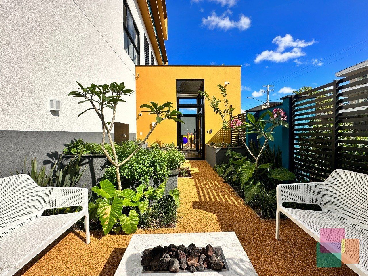 A modern outdoor patio with white metal chairs, a central table with stones, and a yellow building in the background.