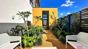 A modern outdoor patio with white metal chairs, a central table with stones, and a yellow building in the background.