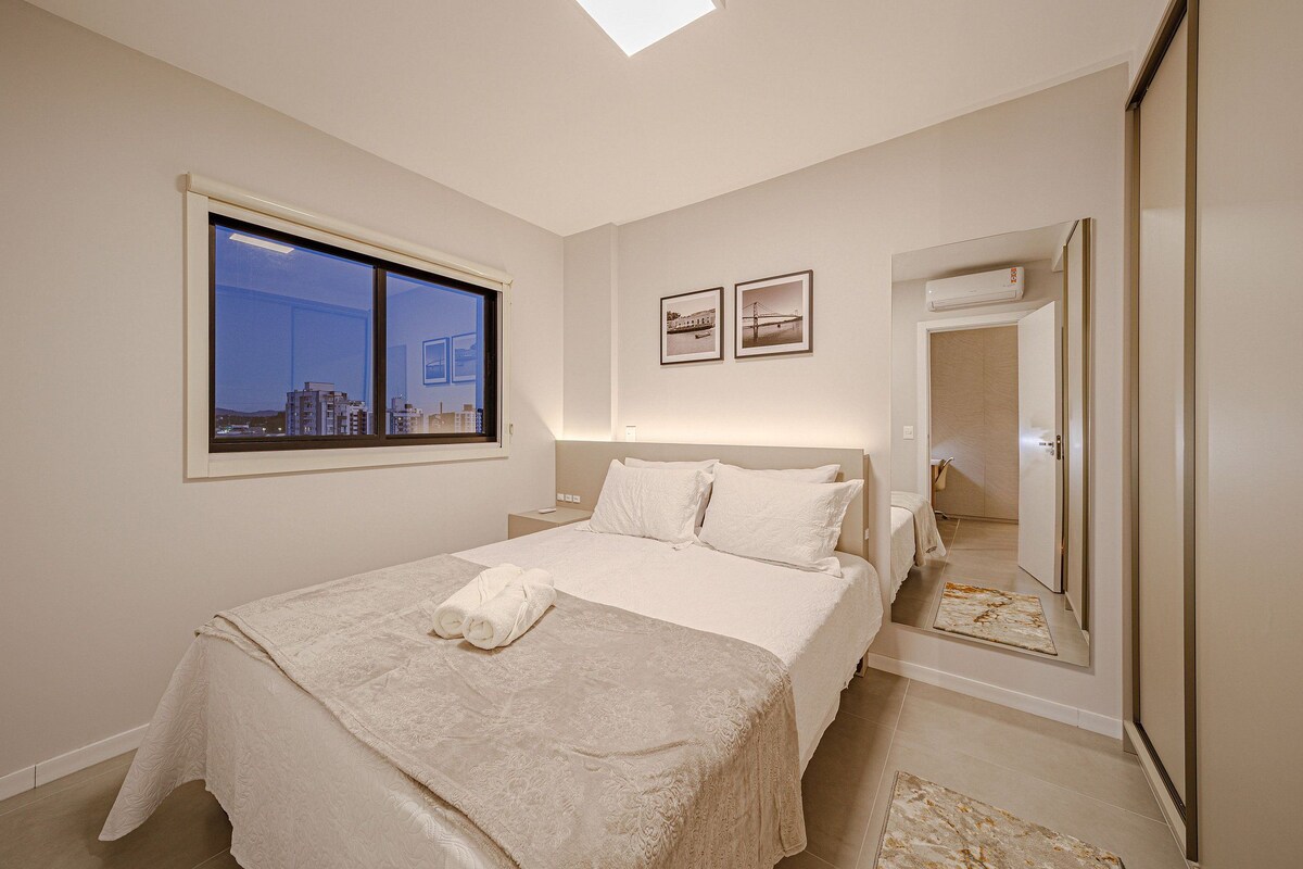 A bedroom with a large window, a bed with a white bedspread, two framed pictures on the wall, and a mirror.