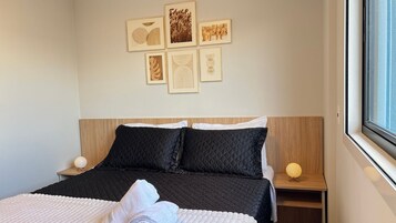 Uma cama bem arrumada com cobertura acolchoada, uma mesa de cabeceira com abajur e uma parede com quadros.
