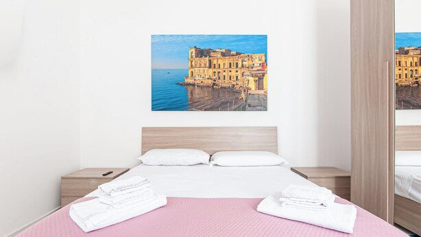 A hotel room with a bed, a pink bedspread, white towels, a wooden headboard, and a cityscape view on the wall. - Apartment in Marechiaro Int 13 (Napoli)