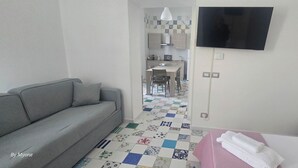 A hotel room with a grey sofa, a bed with a pink bedspread, a TV mounted on the wall, and a kitchen area visible through an open door. - Apartment in Marechiaro Int 13 (Napoli)