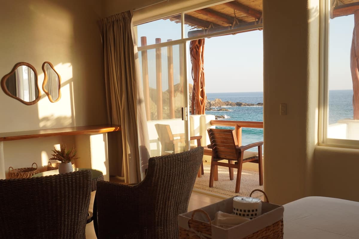 Una habitación con cama, dos sillones de mimbre, una mesa, un ventanal con vista al mar y un espejo en la pared.