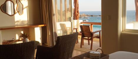 Una habitación con cama, dos sillones de mimbre, una mesa, un ventanal con vista al mar y un espejo en la pared.