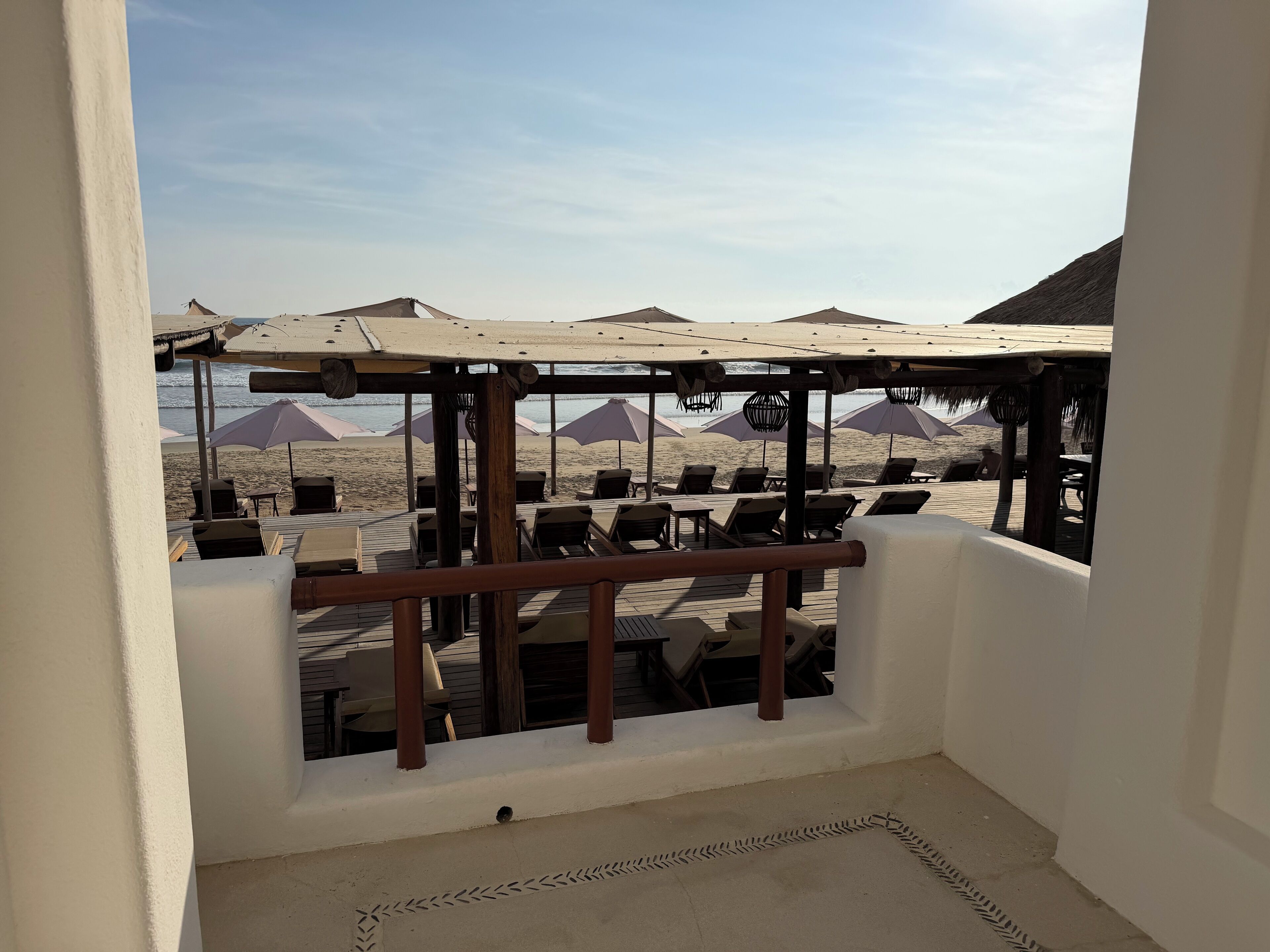 A beachside terrace with wooden tables and chairs, umbrellas, and a view of the beach and ocean.