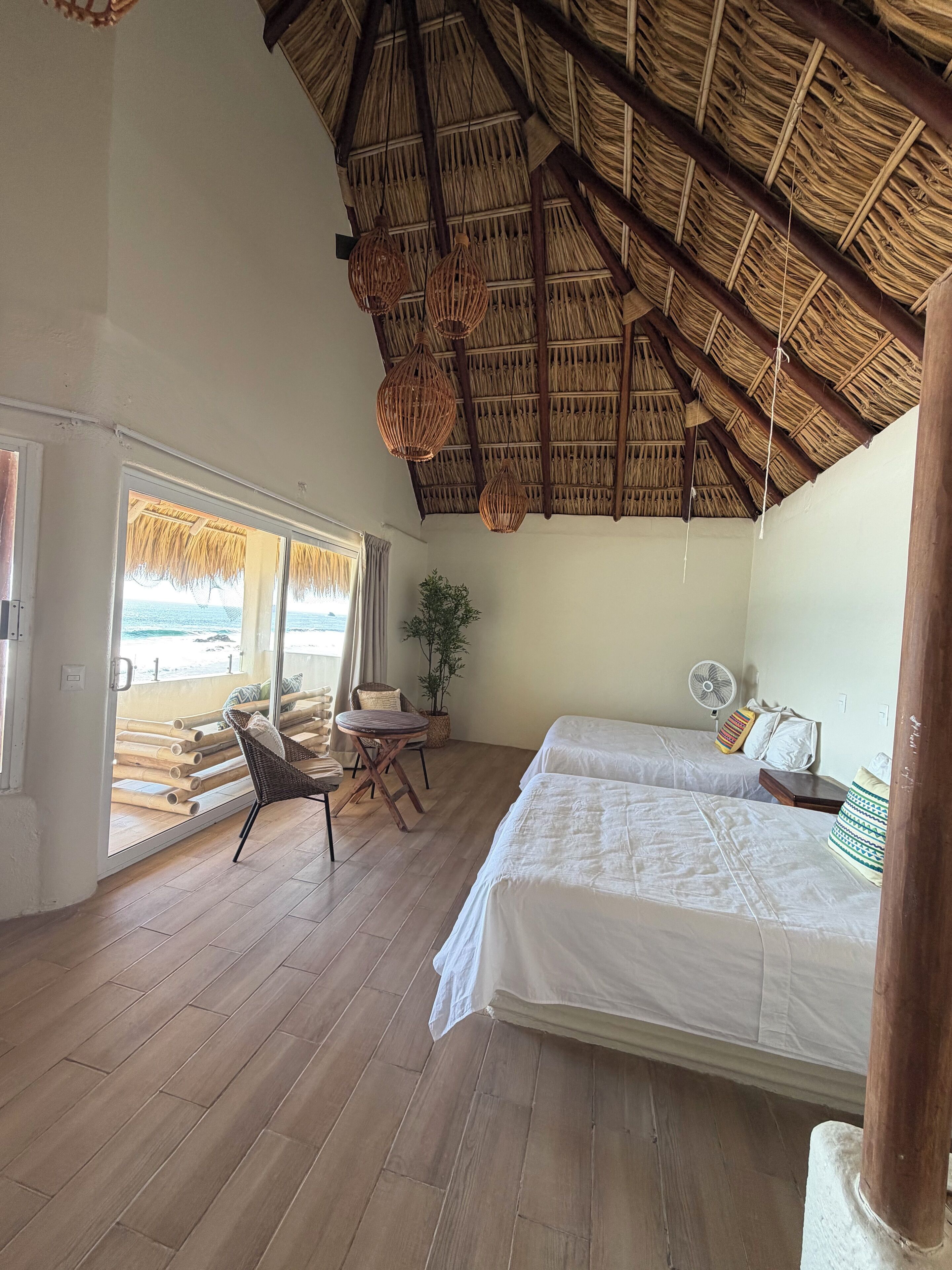 A room with a thatched roof, wooden flooring, two beds, a chair, a fan, and a view of the beach.