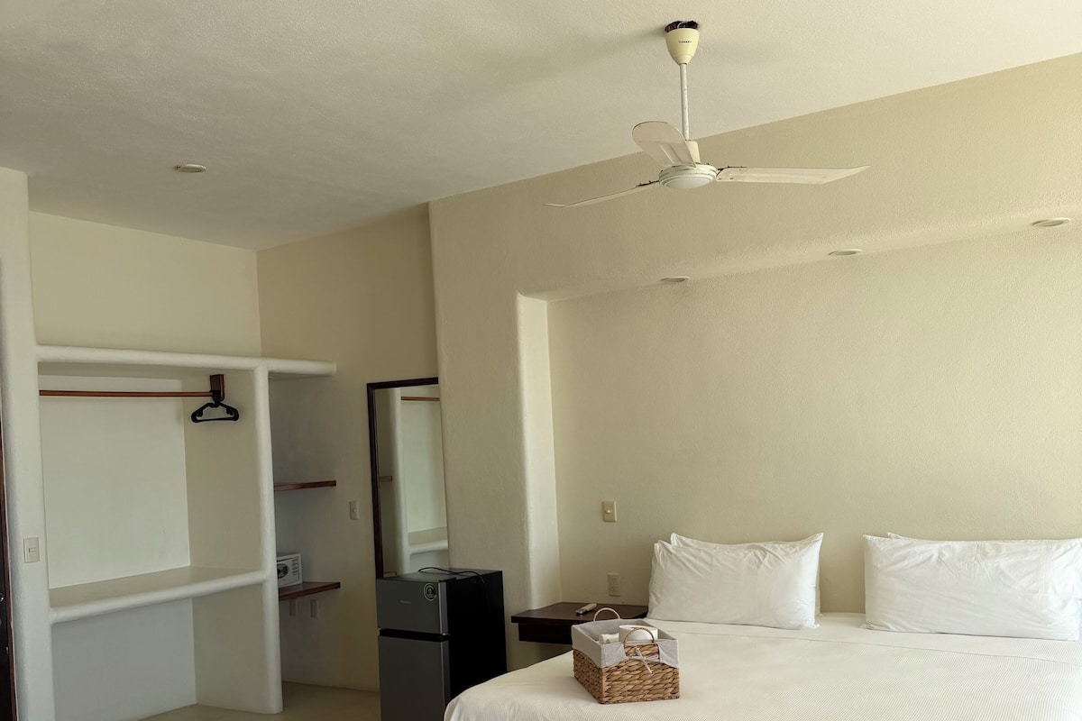 Habitación de hotel con cama, armario, escritorio, silla, espejo y ventilador de techo.