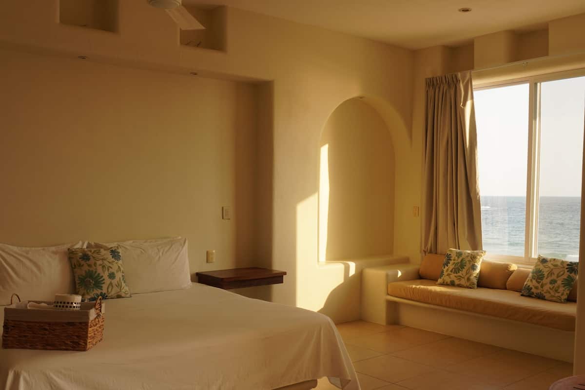 Habitación de hotel con cama, una mesita, una silla, ventana con vista al mar y un ventilador de techo.