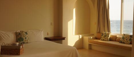 A hotel room with a bed, a small table, a chair, a window with a view of the sea, and a ceiling fan.
