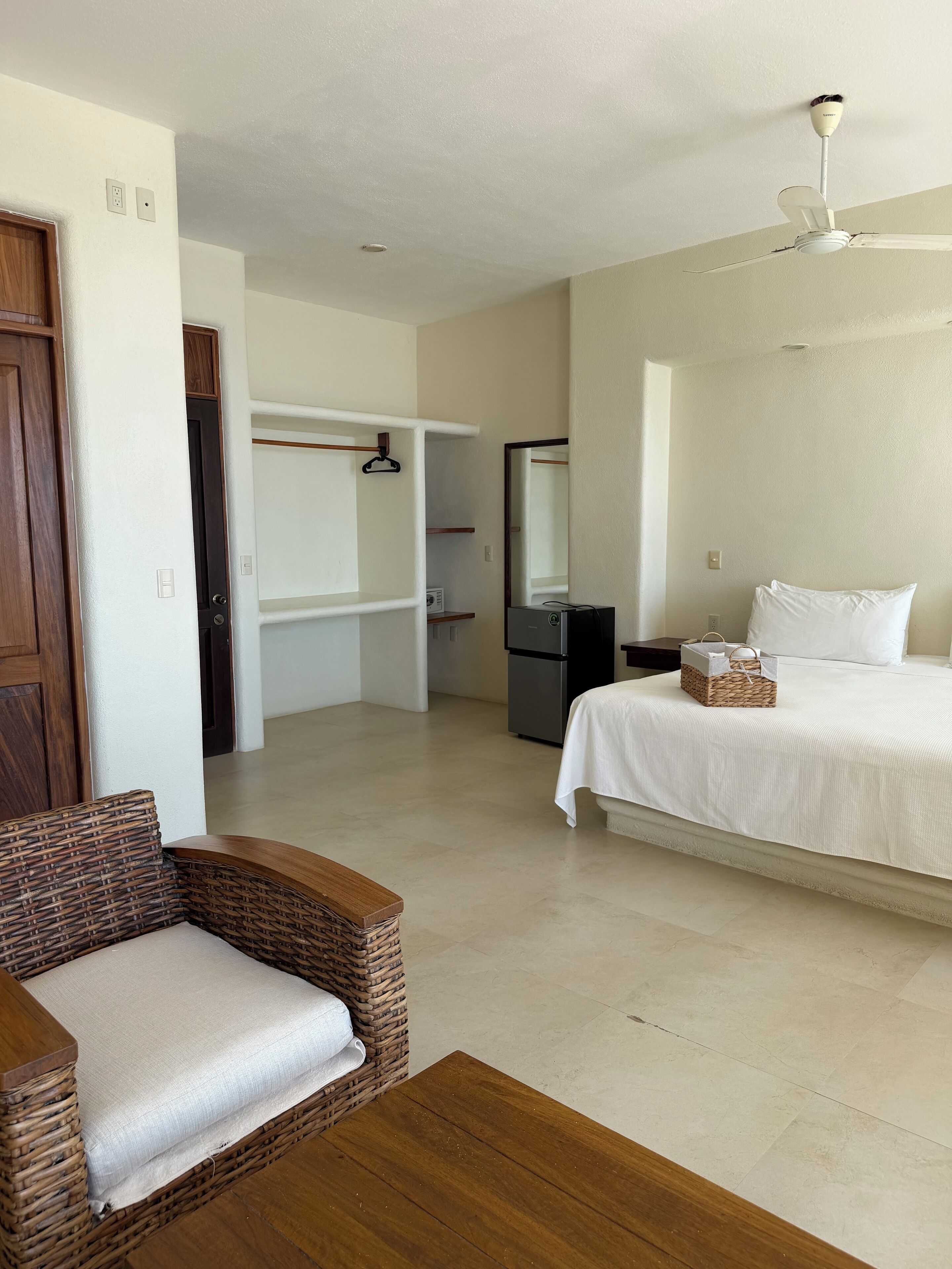 A hotel room with a bed, a wicker chair, a desk, a wardrobe, a mirror, and a ceiling fan.